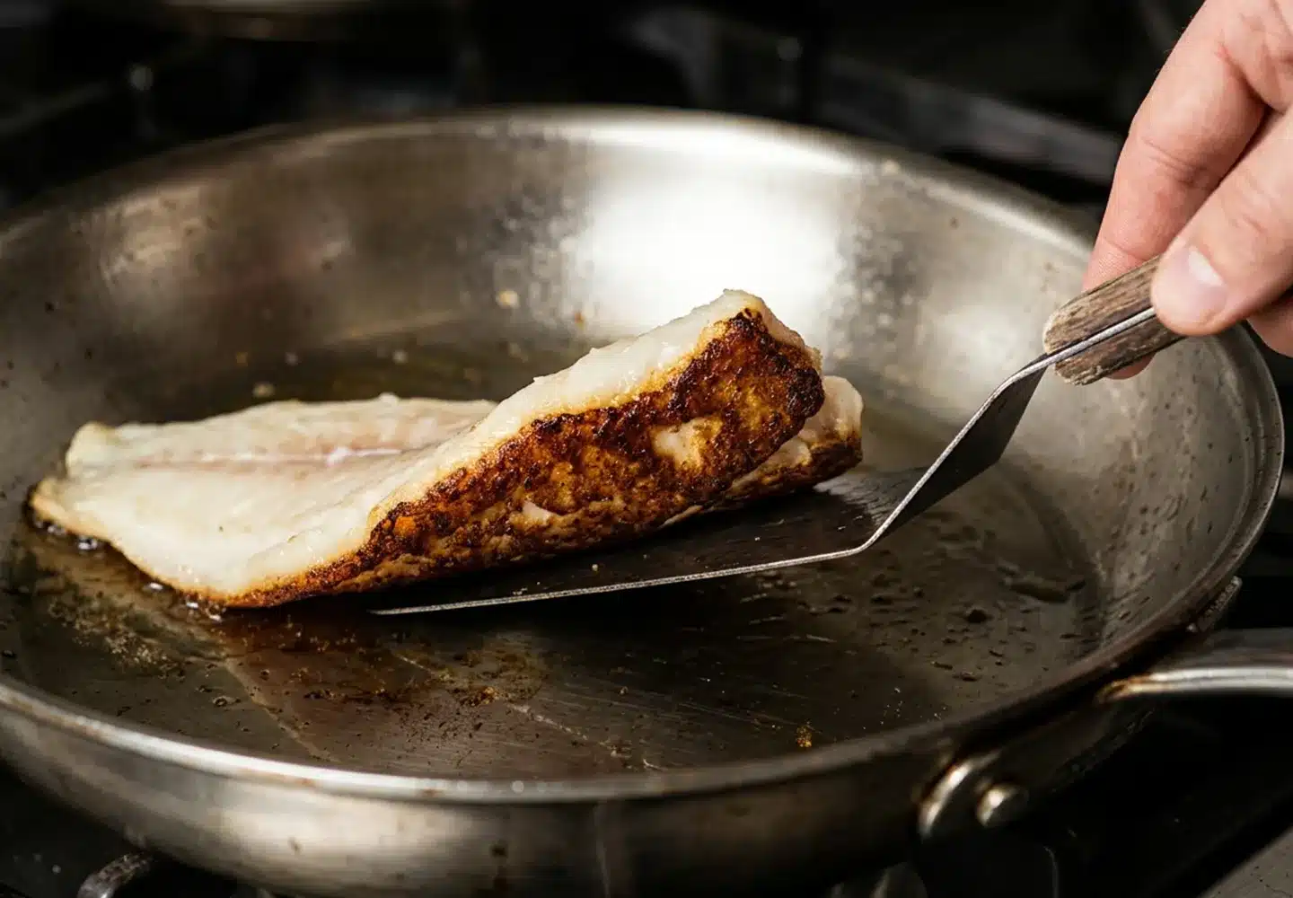 A thin fish spatula lifting the edge of a haddock fillet in a steel pan, showing a dark brown crust underneath.