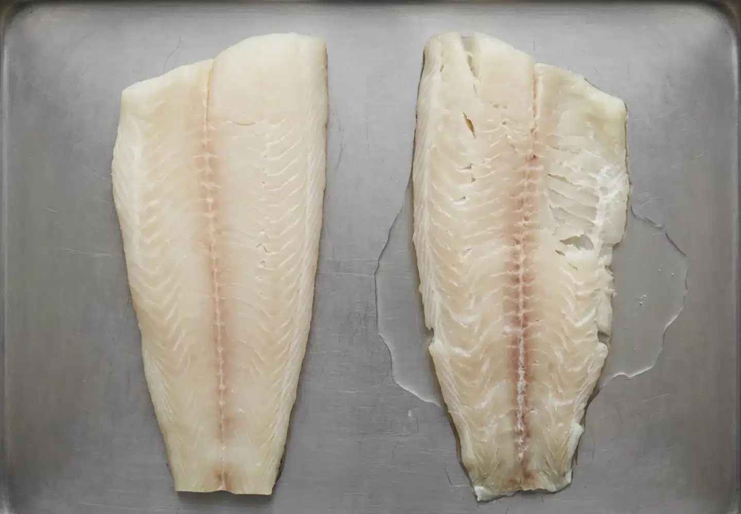 Two raw white fish fillets on a steel tray, one with tight aligned flakes and one visibly looser with liquid pooling.